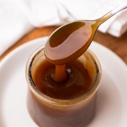 A spoon dripping thick maple caramel sauce into a small jar set on a white plate.