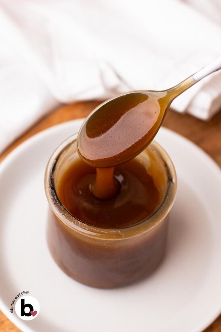 A spoon dripping thick maple caramel sauce into a small jar set on a white plate.