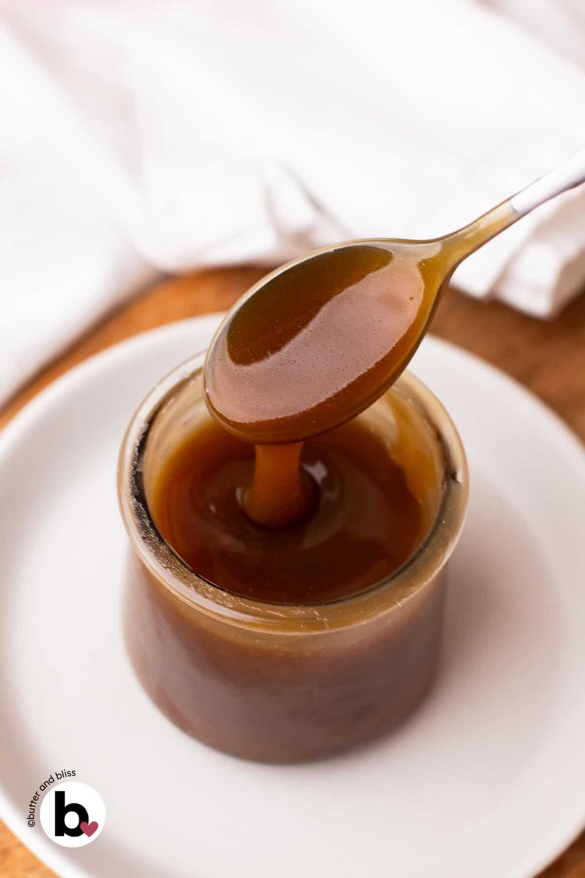 A spoon dripping thick maple caramel sauce into a small jar set on a white plate.