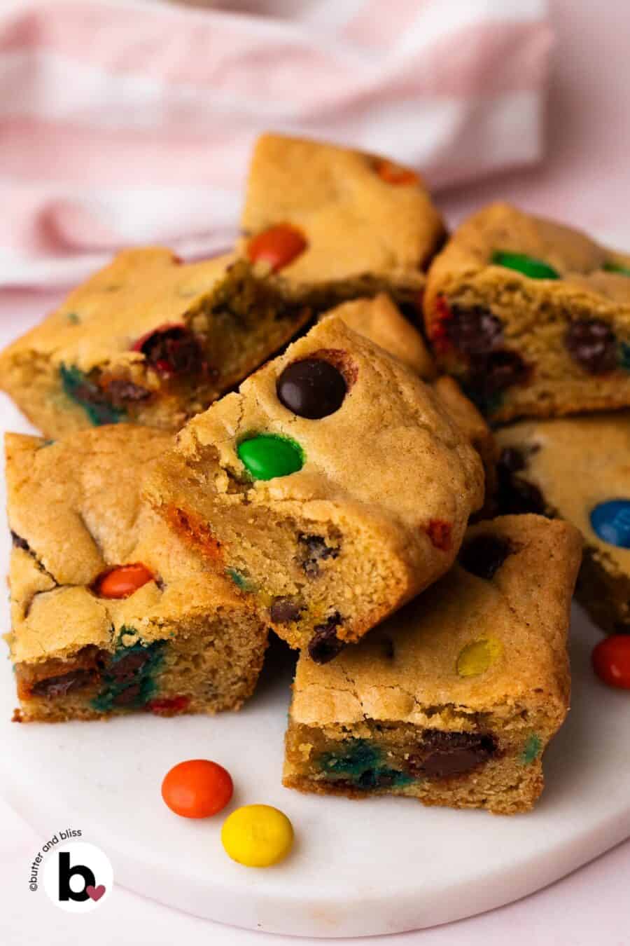 M&M cookie bars squares piled on a white plate next to a pink napkin.