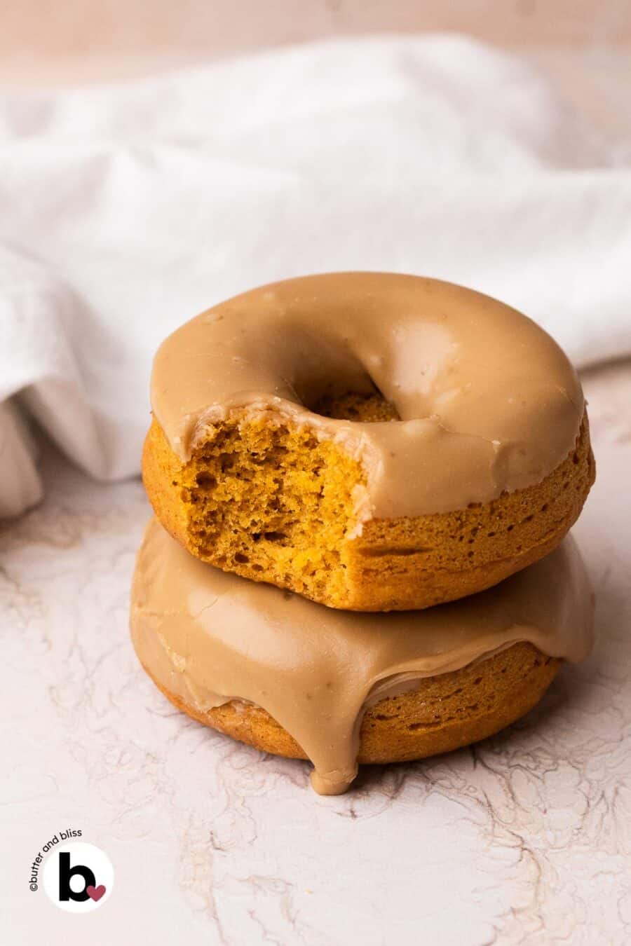 Two maple glazed baked pumpkin donuts stacke on a table with a bite out of one.