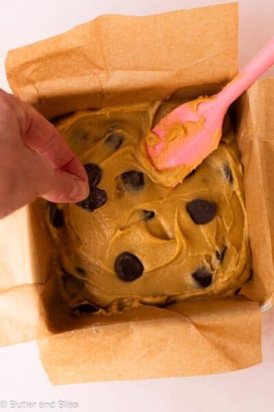 Blondie batter in a square pan with large chocolate chunks, and a hand pressing extra chocolate on top.