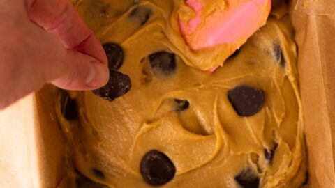 Blondie batter in a square pan with large chocolate chunks, and a hand pressing extra chocolate on top.