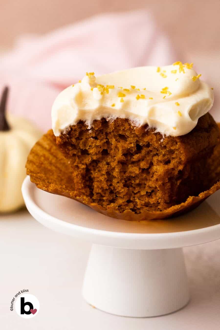 Single pumpkin cupcake with cream cheese frosting with a bite taken to reveal moist center.