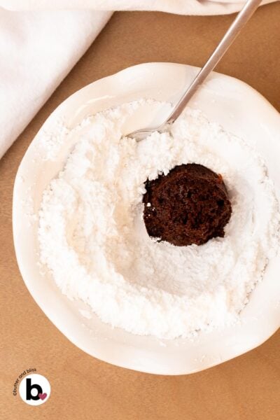 Fudgy chocolate cookie dough ball being rolled through powdered sugar in a bowl.