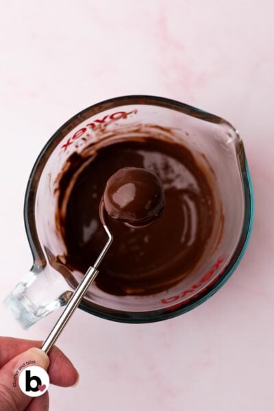 Hand holding a chocolate dipped cookie dough bite over a bowl of melted chocolate.