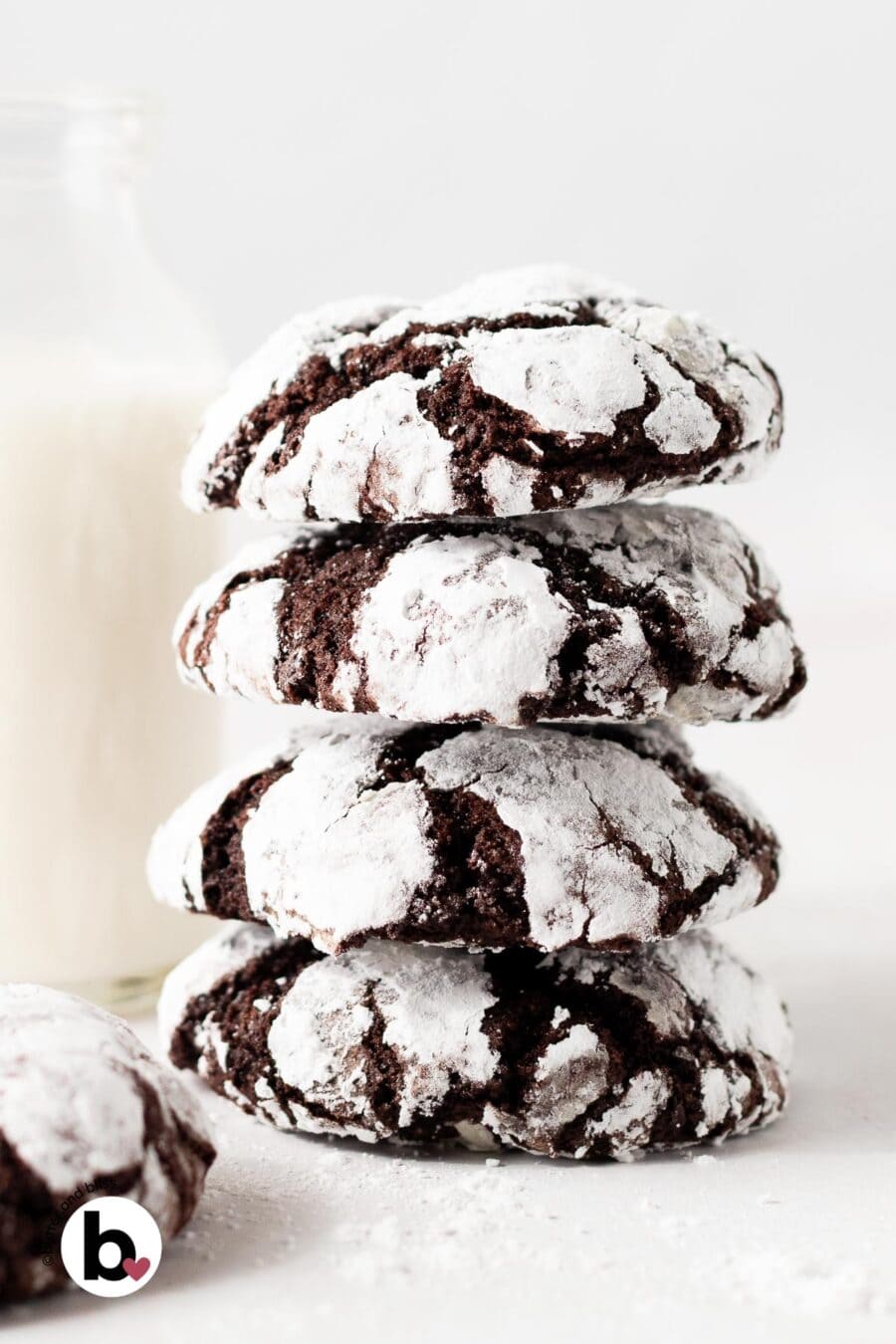 A stack of bright white chocolate crinkle cookies on a table next to a glass of milk.